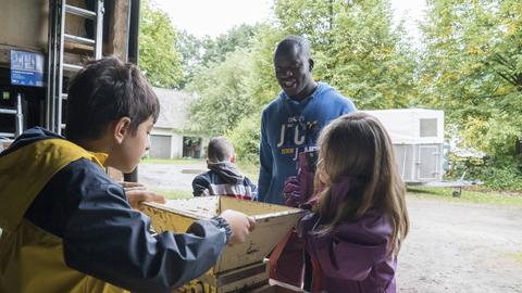 Brighton Paddey Chimanyiwa arbeitet auf dem Schulbauerhof in Bielefeld-Ummeln mit Kindern an der Saftpresse. Im Hintergrund steht ein Pferdeanhänger auf dem Hof, dahinter sind Bäume und Gebüsch zu sehen.