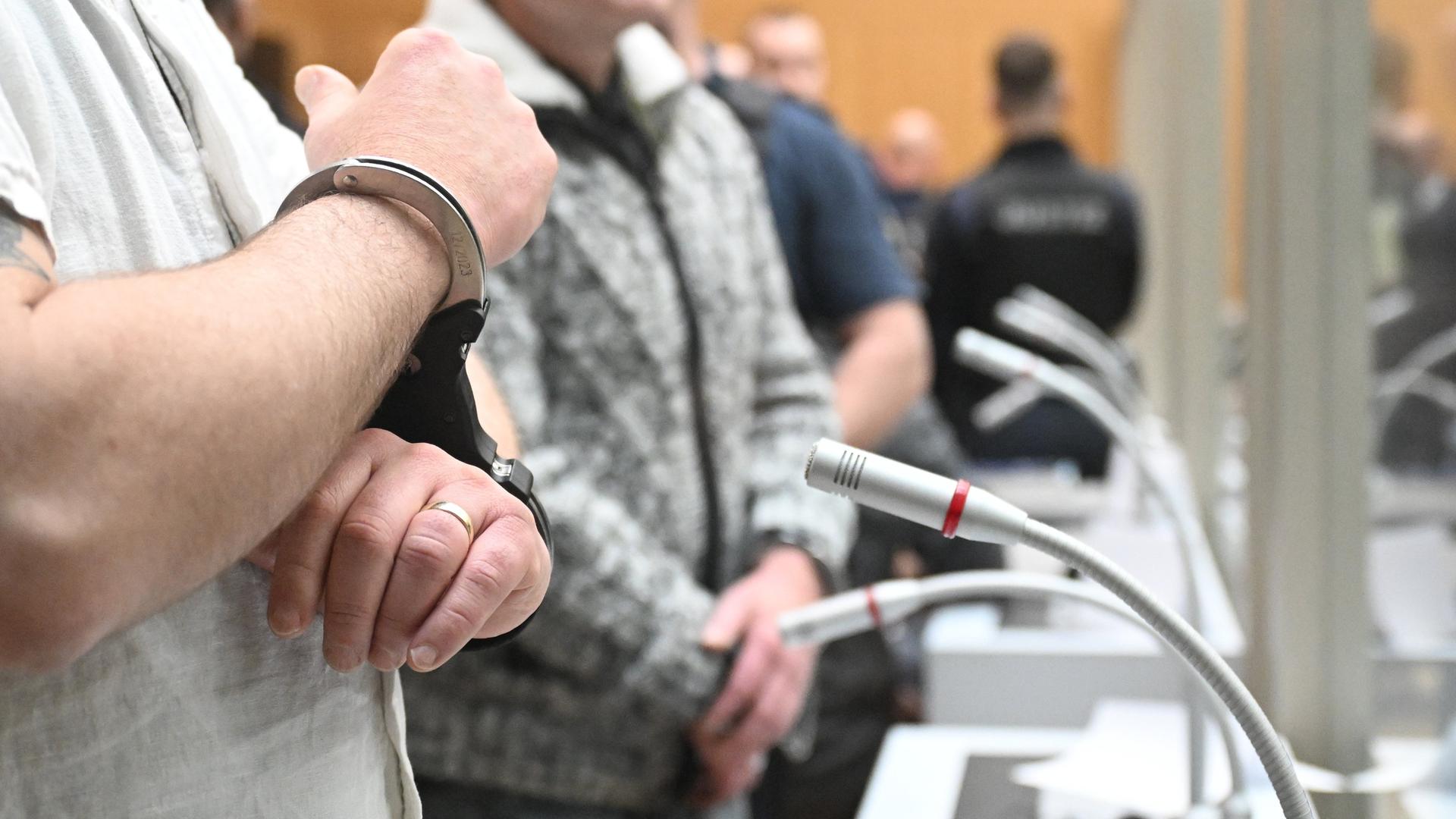 Angeklagte stehen in Hand-Schellen beim beim Beginn eines Prozesses um "Reichs-Bürger", die mutmaßlich einen Umsturz in Deutschland geplant haben sollen, im Gerichts-Saal an Mikrofonen. 