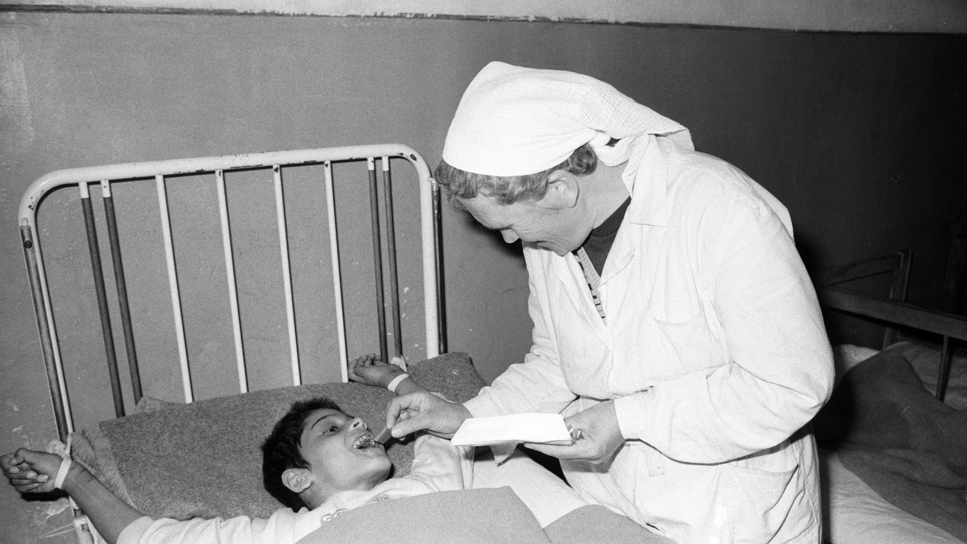 Eine Pflegerin füttert auf dem Schwarzweißbild von 1990 einen an ein Bettgestell gefesselten Jungen in einem Heim in Badacin, Rumänien.