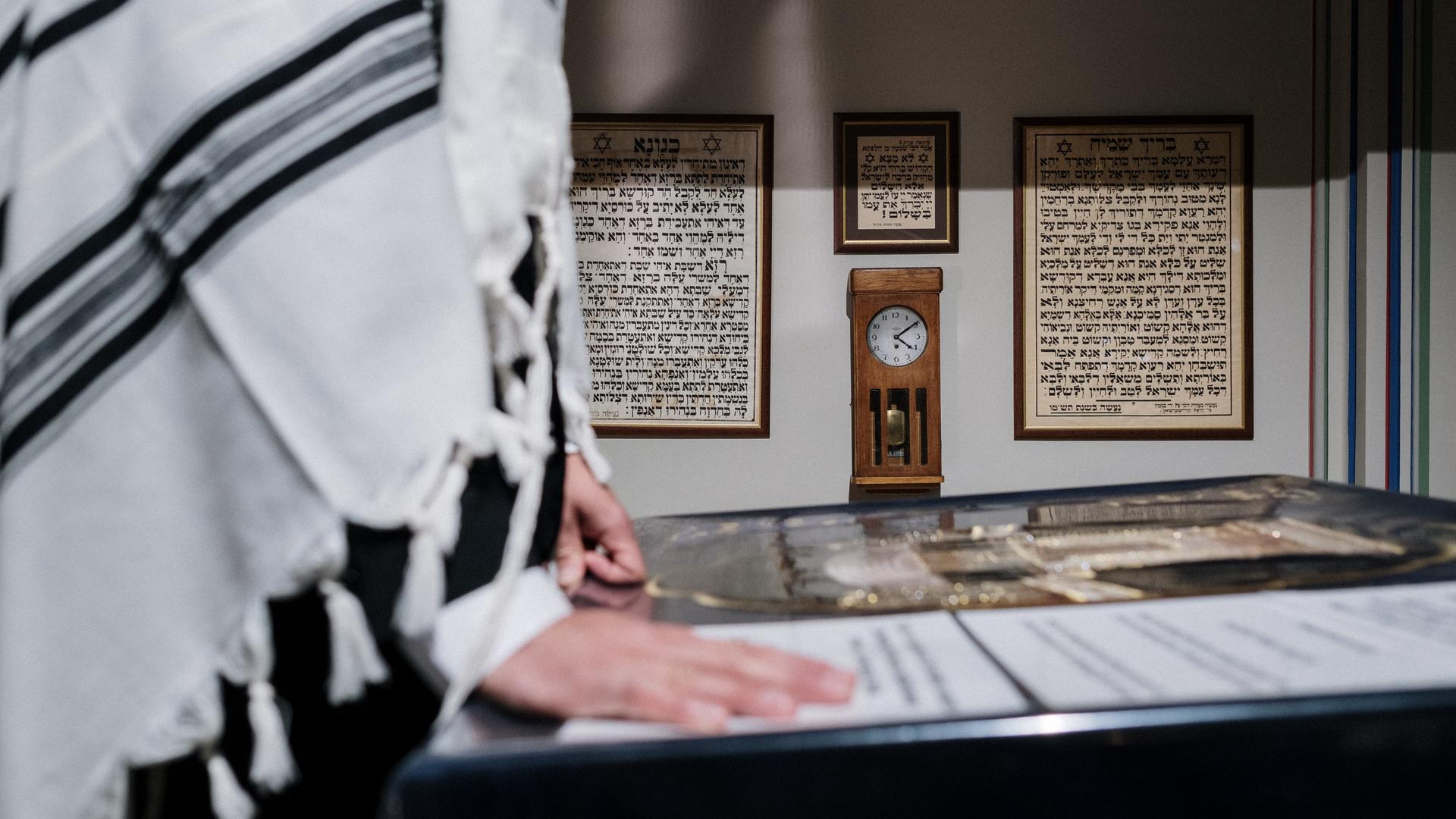 Eine Person mit einem über die Schultern geworfenen Tallit steht vor einer hebräischen Schrift.