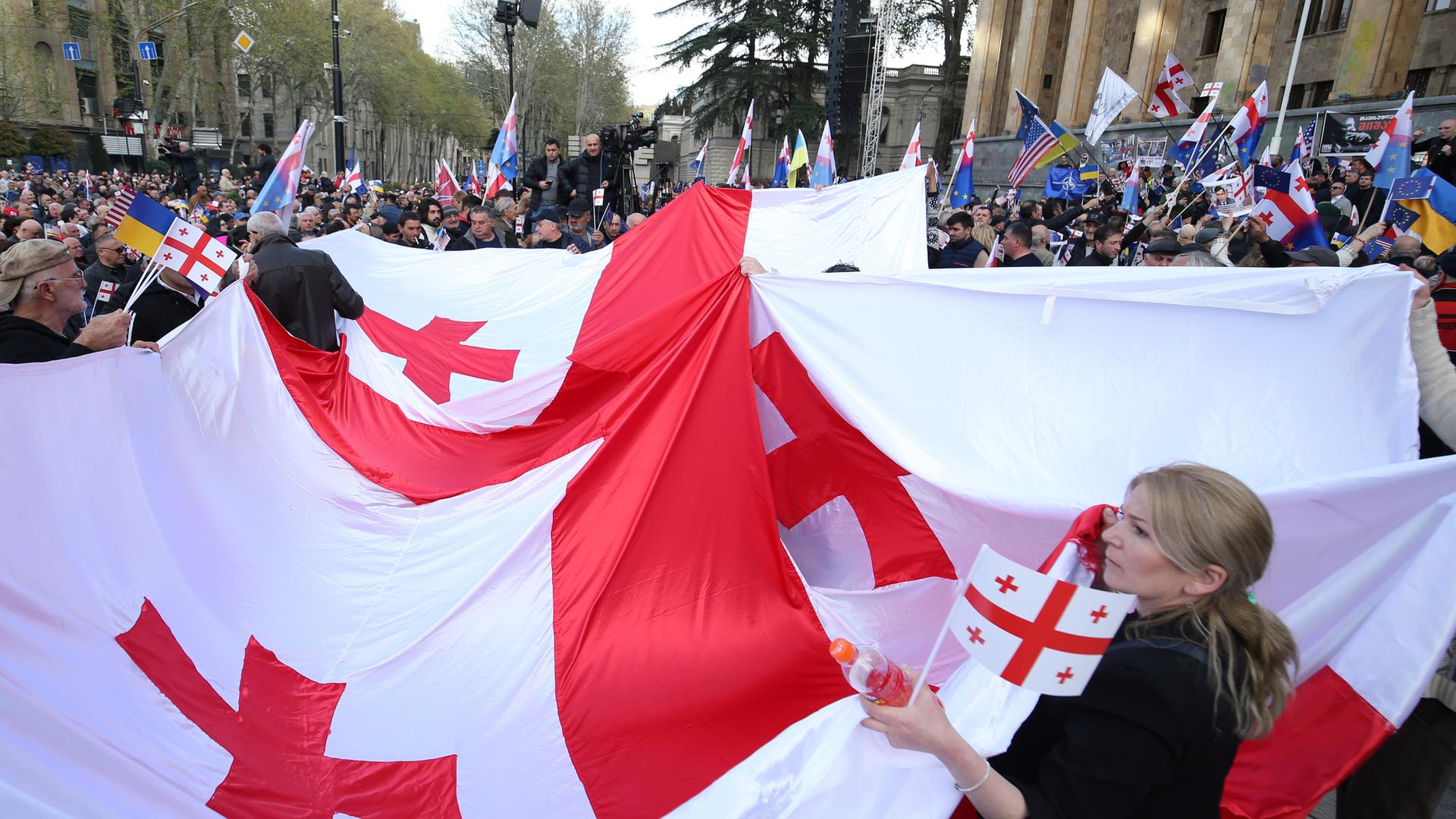 Demonstranten halten eine große georgische Fahne während eines Protests hoch. Im Hintergrund sind viele andere Protestierende zu sehen, die auch Flaggen der EU und der Ukraine hochhalten.