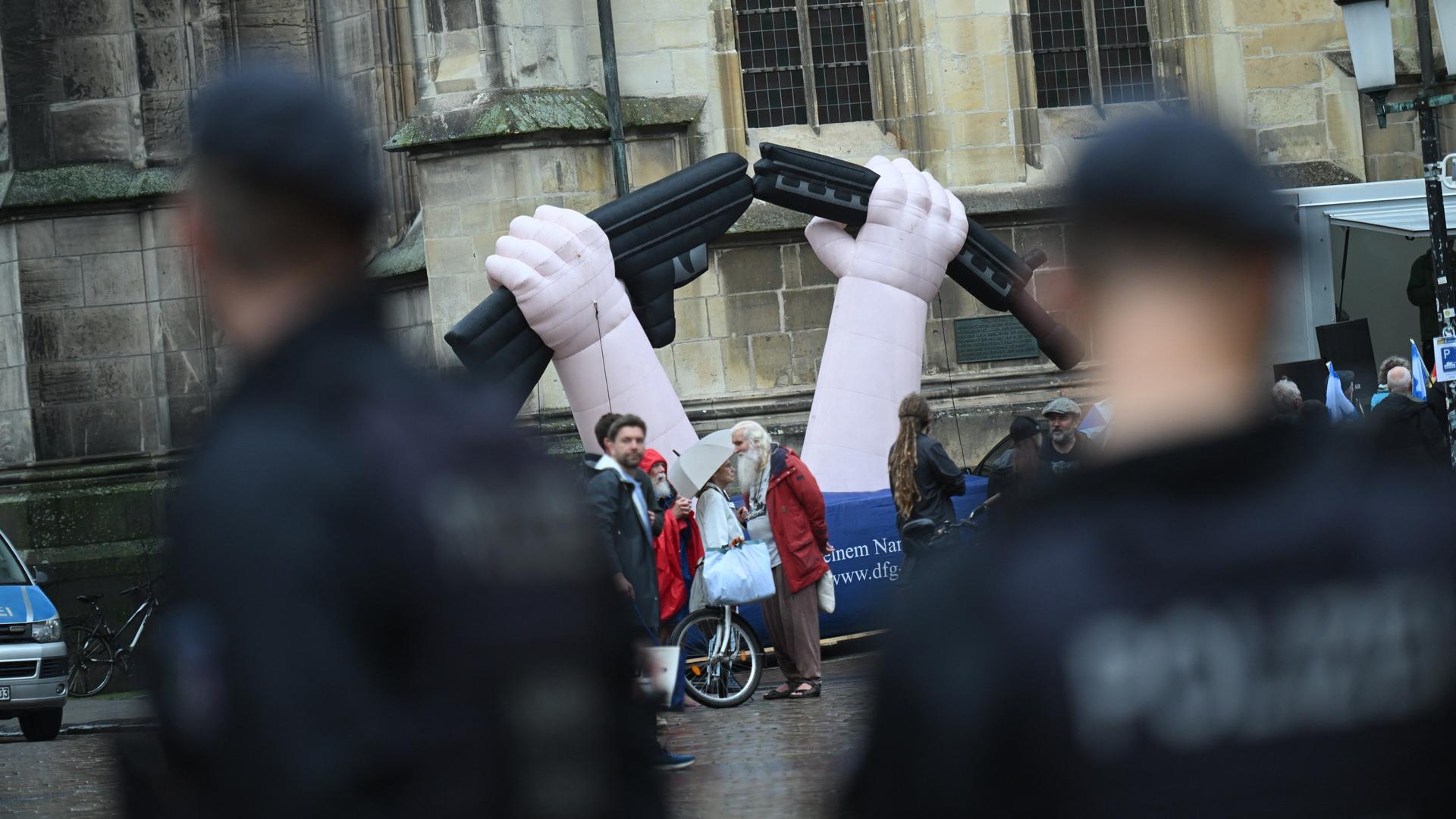 Vor der Kirche steht eine aufblasbare Skulptur mit zwei Händen, die ein Gewehr zerbrechen. Daneben Demonstranten. Im Vordergrund unscharf zwei Polizisten. 