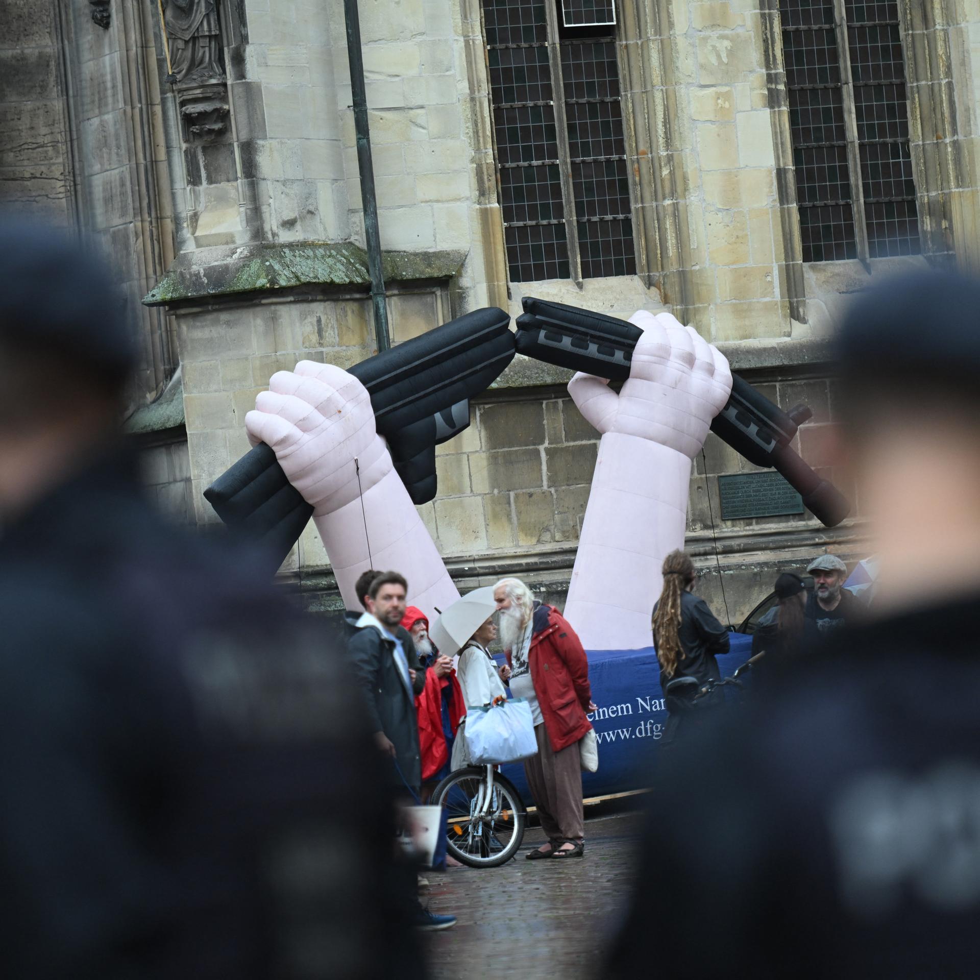 Vor der Kirche steht eine aufblasbare Skulptur mit zwei Händen, die ein Gewehr zerbrechen. Daneben Demonstranten. Im Vordergrund unscharf zwei Polizisten. 