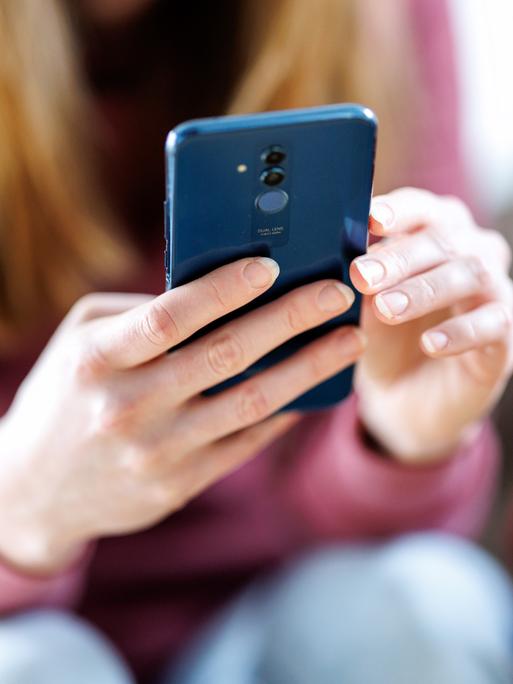 Eine junge Frau sitzt und hält ein Smartphone mit beiden Händen. Sie blickt auf das Display. Ihr Gesicht ist unscharf im Hintergrund. Eine junge Frau sitzt und hält ein Smartphone mit beiden Händen. Sie blickt auf das Display. Ihr Gesicht ist unscharf im Hintergrund.