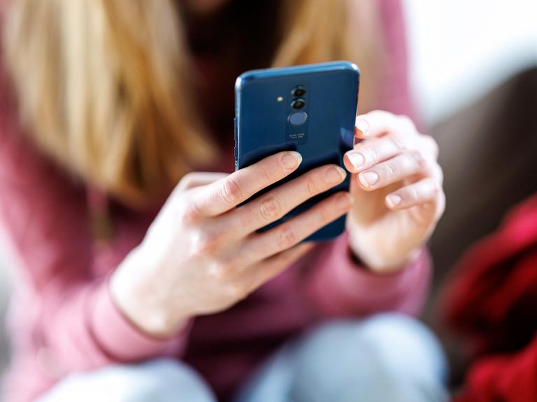Eine junge Frau sitzt und hält ein Smartphone mit beiden Händen. Sie blickt auf das Display. Ihr Gesicht ist unscharf im Hintergrund.