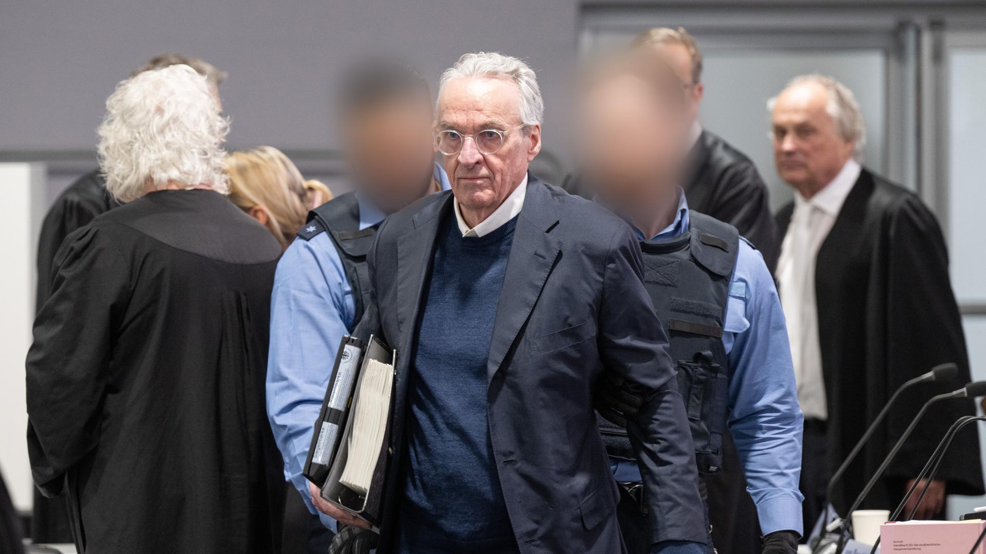 Heinrich Prinz Reuß wird von zwei Polizisten in den Gerichtssaal gebracht. Im Hintergrund stehen mehrere Anwälte in schwarzen Roben. 