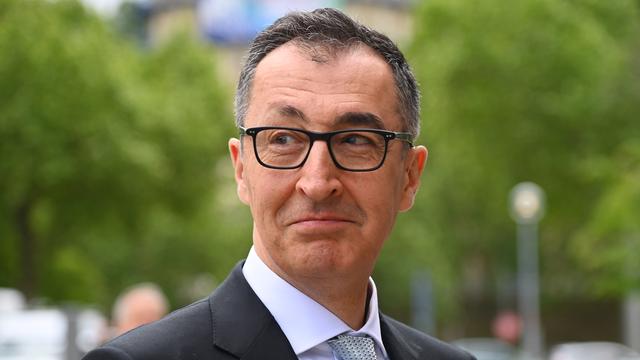 Cem Özdemir trägt eine Brille mit schwarzem Rahmen und lächelt. Im Hintergrund sind grüne Bäume.