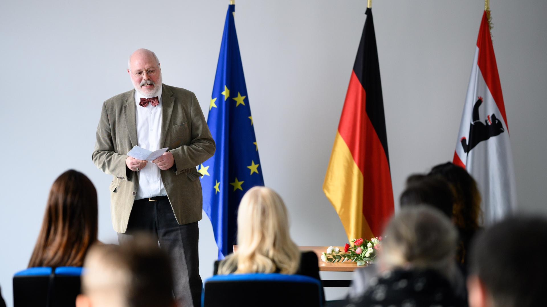 Ein Mann steht neben einer europäischen, einer deutschen und einer Berliner Flagge. Vor ihm sitzen mehrere Menschen auf Stühlen.