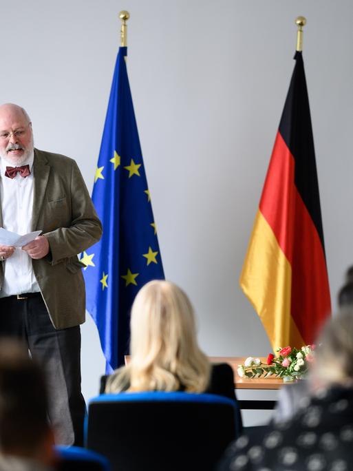 Ein Mann steht neben einer europäischen, einer deutschen und einer Berliner Flagge. Vor ihm sitzen mehrere Menschen auf Stühlen.