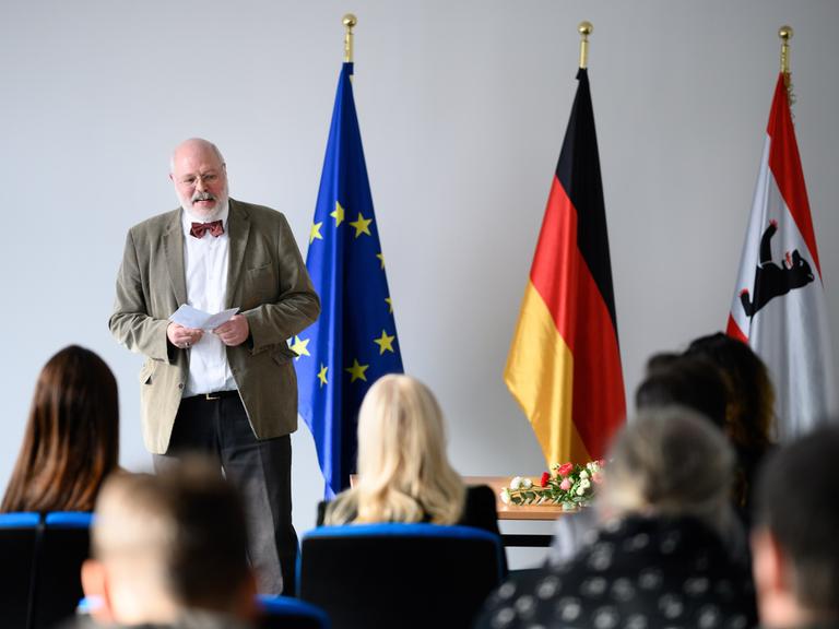 Ein Mann steht neben einer europäischen, einer deutschen und einer Berliner Flagge. Vor ihm sitzen mehrere Menschen auf Stühlen.