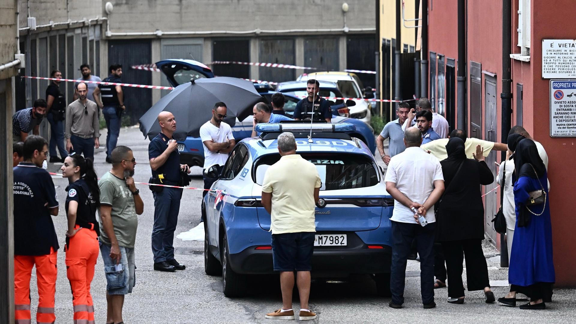 Ein Polizeiwagen steht vor Absperrbändern in einer Gasse. Viele Menschen und mehrere Beamte stehen darum herum. Ein Polizeiwagen steht vor Absperrbändern in einer Gasse. Viele Menschen und mehrere Beamte stehen darum herum.