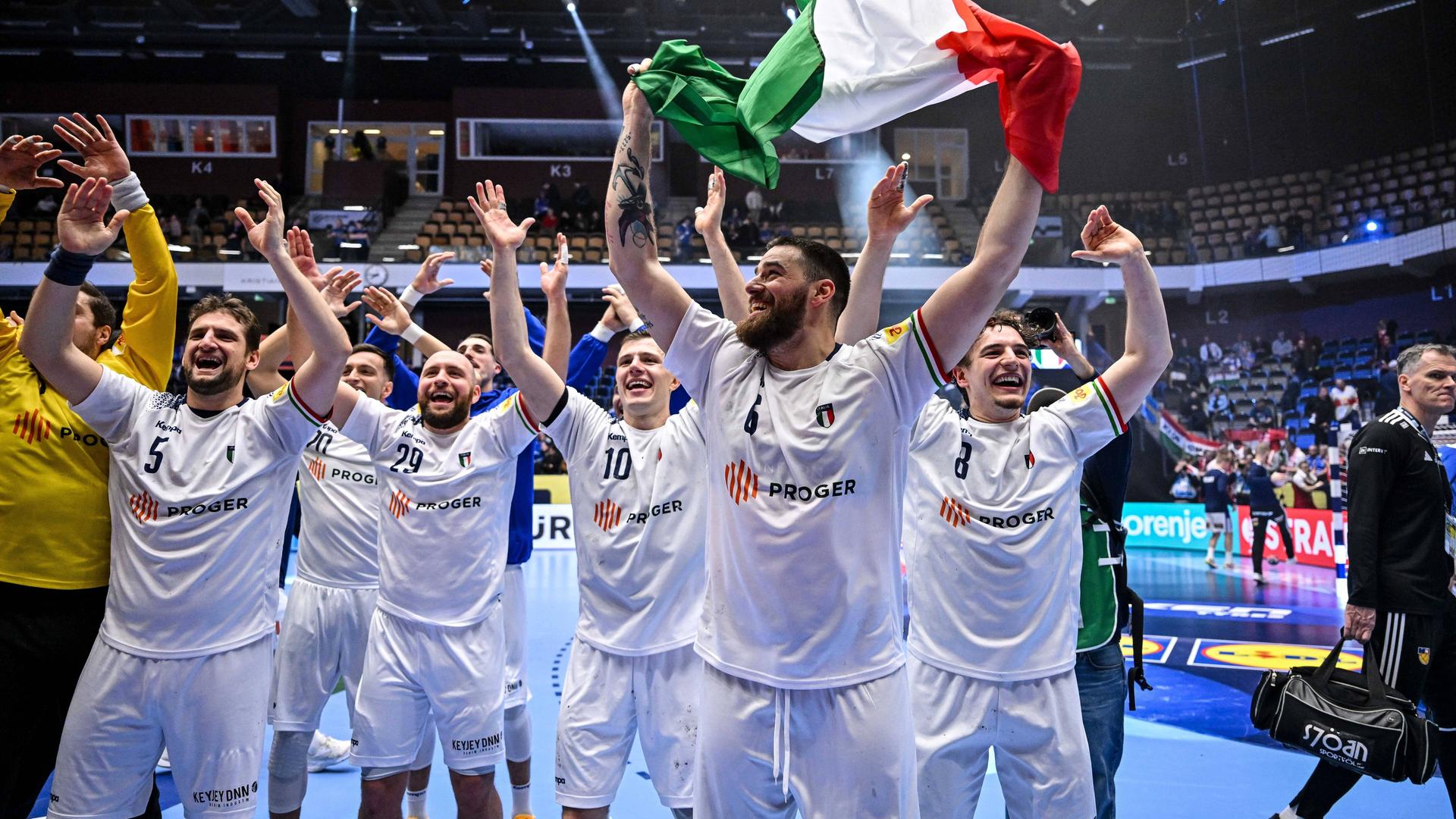 Italiens Handballer reißen ihre Arme hoch und feiern ihren EM-Sieg gegen Polen. Einer der Spieler jubelt mit einer italienischen Flagge in den Händen.