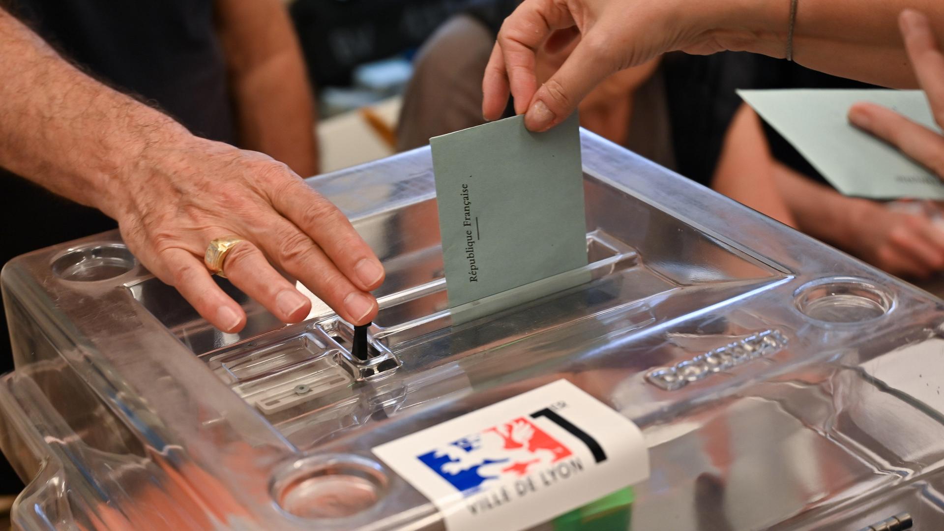 Es ist eine Wahlurne zu sehen sowie zwei Hände. Eine Hand öffnet die Wahlurne, die andere Hand wirft einen geschlossenen Stimmzettel ein.