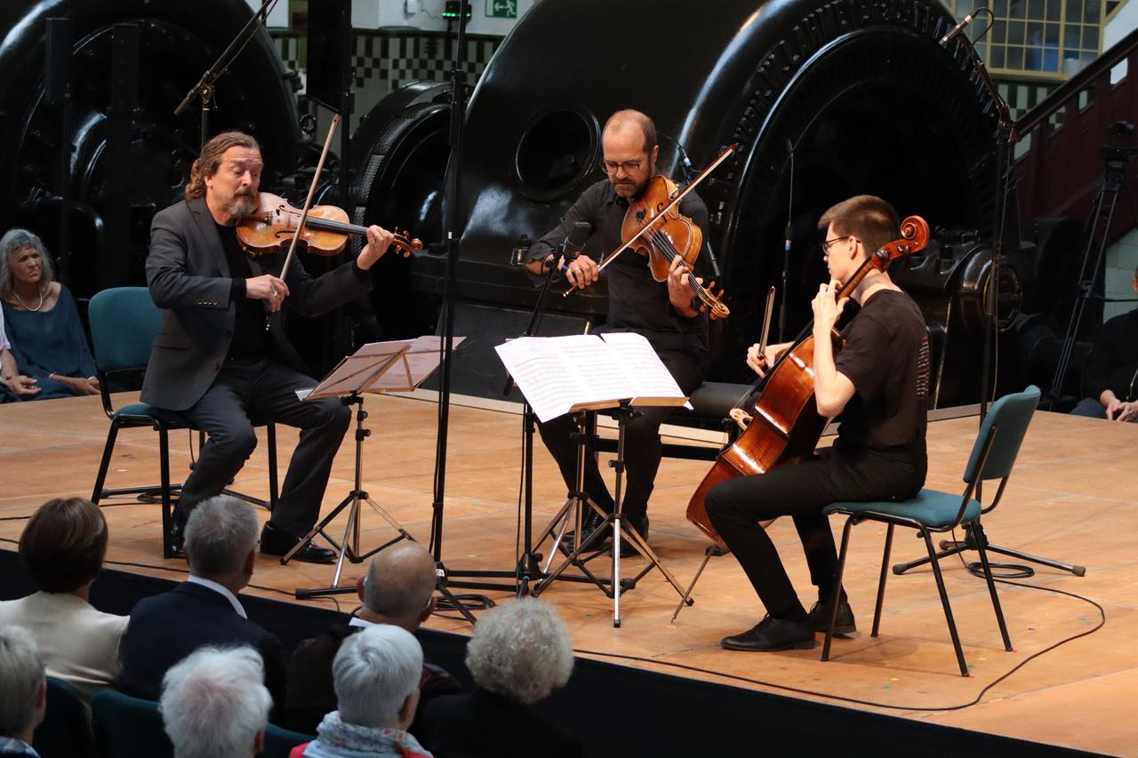 Drei schwarz gekleidete Musiker mit Streichinstrumenten sitzen auf einer Bühne. Im Hintergrund historische Turbinen, verschwommen im Vordergrund die ersten Reihen des Publikums. Drei schwarz gekleidete Musiker mit Streichinstrumenten sitzen auf einer Bühne. Im Hintergrund historische Turbinen, verschwommen im Vordergrund die ersten Reihen des Publikums.