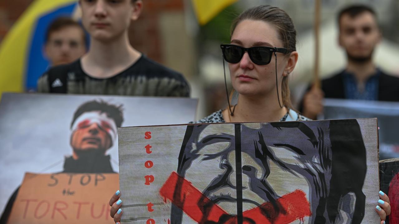 Ukrainische Aktivisten erinnern bei Protesten in Krakau an die ukrainischen Gefangenen in russischer Haft. Eine Frau mit Sonnenbrille hält ein Plakat, das einen schreienden Mann zeigt, dessen Mund rot durchgestrichen ist, mit dem Schriftzug "Stop Torture" (Stoppt Folter) 