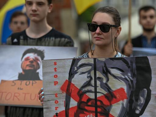 Ukrainische Aktivisten erinnern bei Protesten in Krakau an die ukrainischen Gefangenen in russischer Haft. Eine Frau mit Sonnenbrille hält ein Plakat, das einen schreienden Mann zeigt, dessen Mund rot durchgestrichen ist, mit dem Schriftzug "Stop Torture" (Stoppt Folter) 