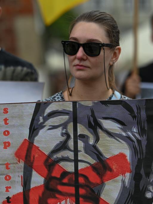 Ukrainische Aktivisten erinnern bei Protesten in Krakau an die ukrainischen Gefangenen in russischer Haft. Eine Frau mit Sonnenbrille hält ein Plakat, das einen schreienden Mann zeigt, dessen Mund rot durchgestrichen ist, mit dem Schriftzug "Stop Torture" (Stoppt Folter) 
