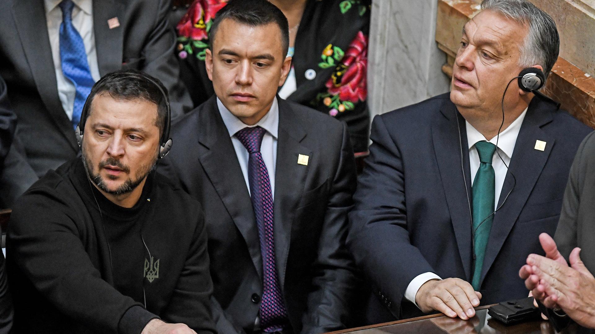 Selenskyj sitzt im Parlament in Buenos Aires an einem Pult, neben ihm unter anderem Orban. Beide tragen Kopfhörer.