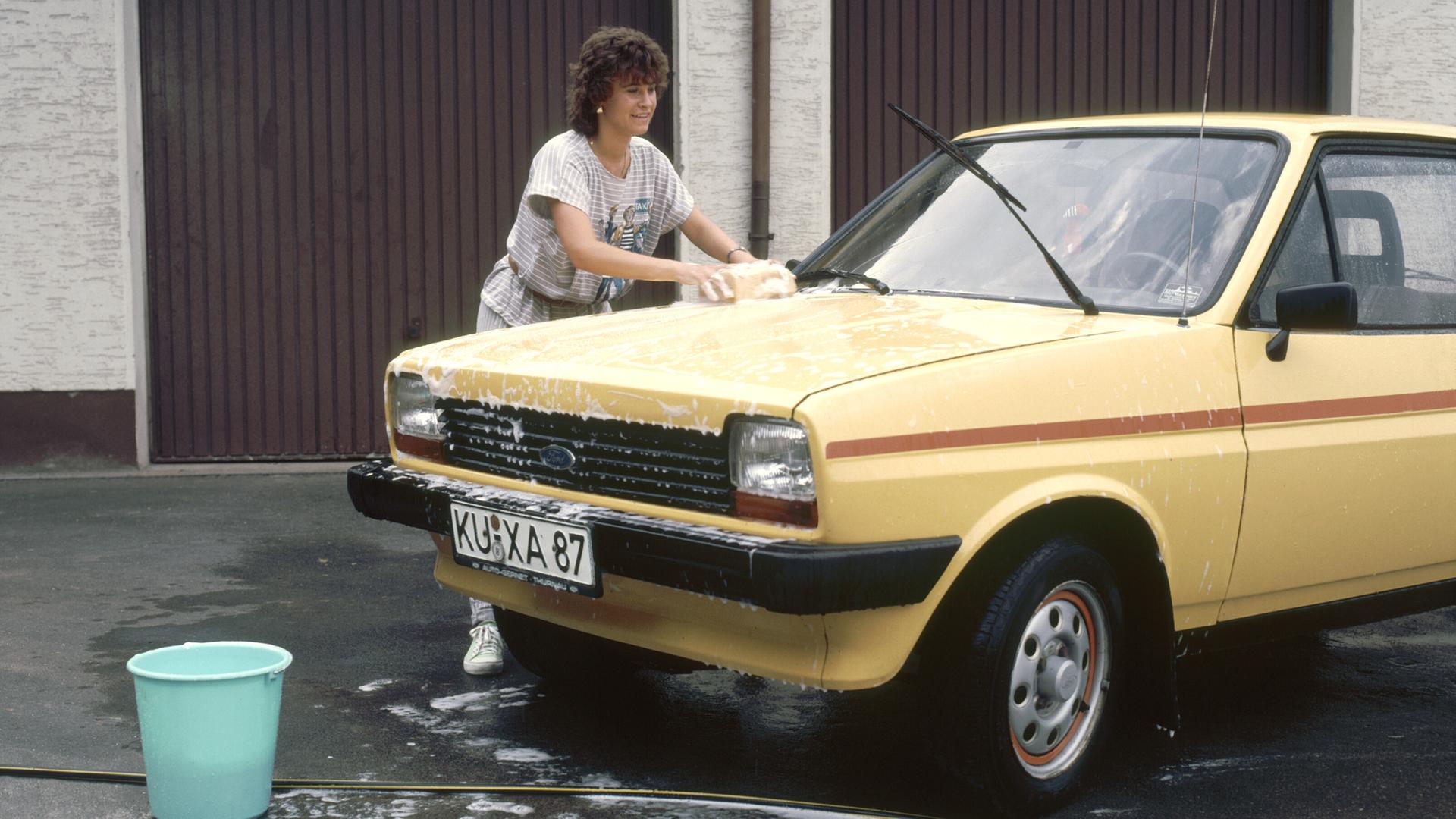 Eine Frau wäscht mit einem Schwamm einen gelben Ford, aufgenommen 1989. Im Hintergrund sind braune Garagentore zu sehen.