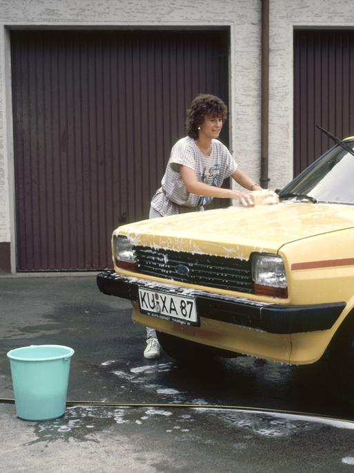 Eine Frau wäscht mit einem Schwamm einen gelben Ford, aufgenommen 1989. Im Hintergrund sind braune Garagentore zu sehen.