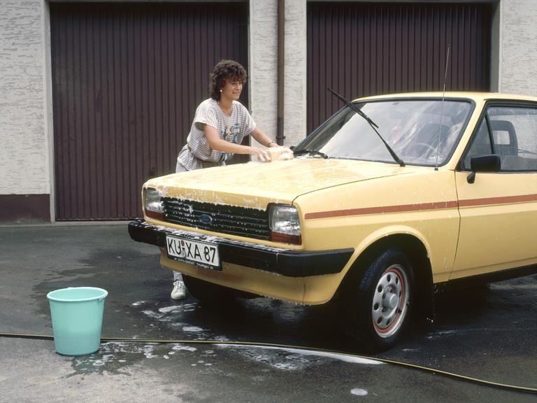 Eine Frau wäscht mit einem Schwamm einen gelben Ford, aufgenommen 1989. Im Hintergrund sind braune Garagentore zu sehen.