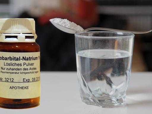 Ein braunes Arzneimittelglas mit Pentobarbital-Natrium, daneben ein gefülltes Wasserglas, auf dem ein Löffel mit dem Pulver liegt. Das tödlich wirkende Mittel wird in der Schweiz beim assistierten Suizid verwendet