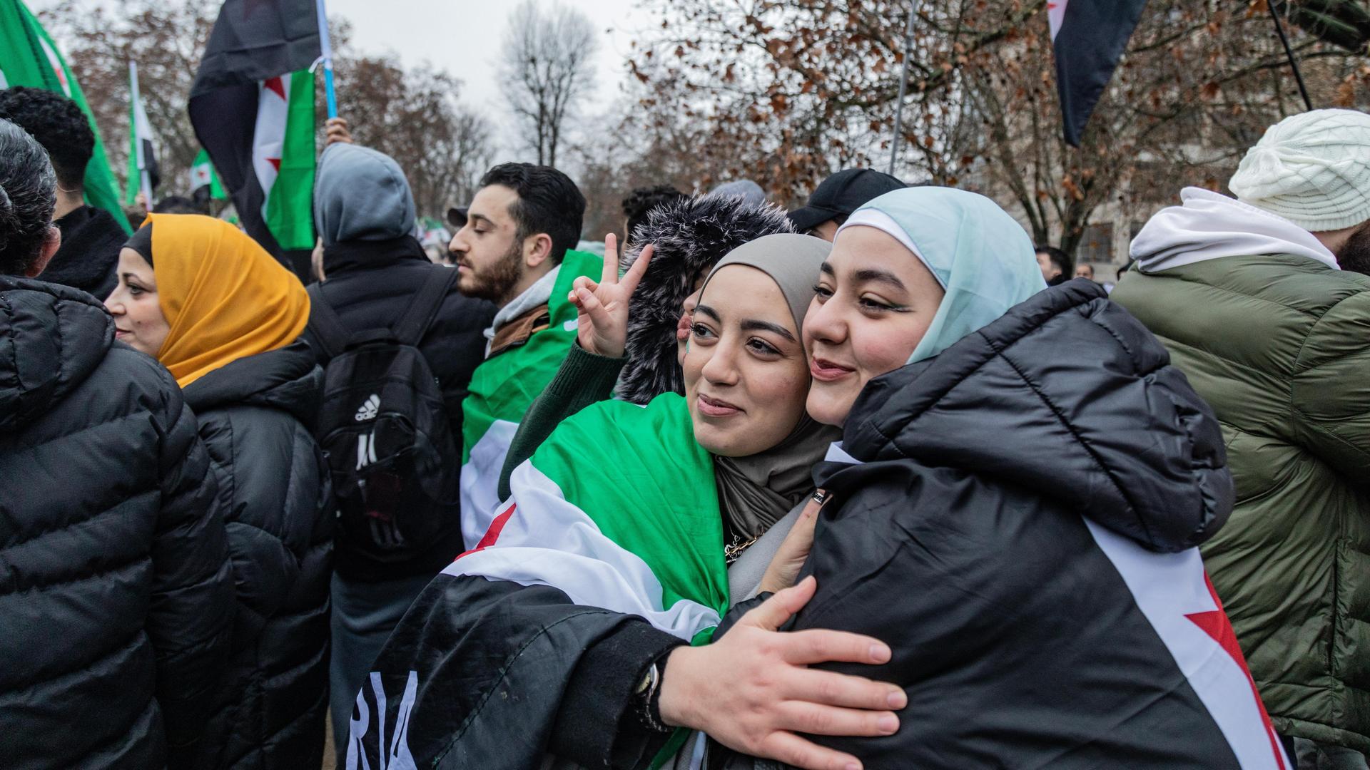 Zwei junge Frauen, teilweise in eine syrische Flagge gehüllt, umarmen sich in einer Menschenmenge in Berlin.