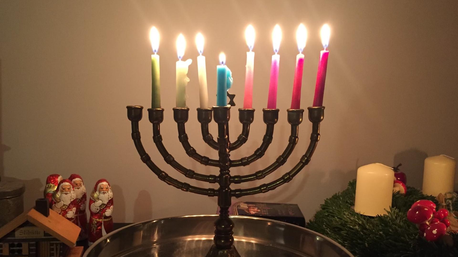 Ein Chanukka-Leuchter mit bunten Kerzen. Im Hintergrund sind Schoko-Weihnachtsmänner zu sehen.