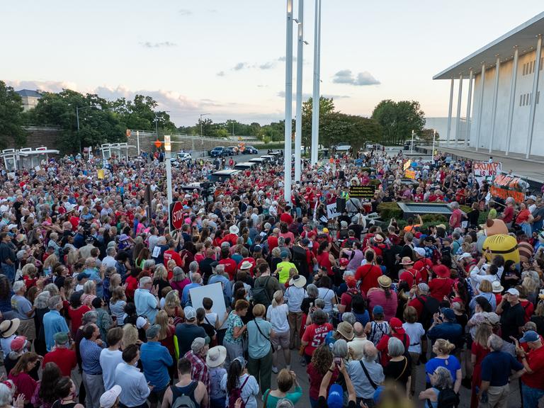 Demonstranten stehen bei Abendlicht vor dem hellen Bau des Kennedy Center. Demonstranten stehen bei Abendlicht vor dem hellen Bau des Kennedy Center.