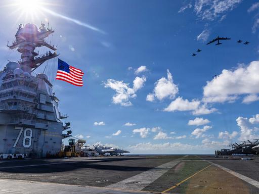 Der größten Flugzeugträger der Welt mit einer US-Flagge vor blauem Himmel und Militärflugzeugen