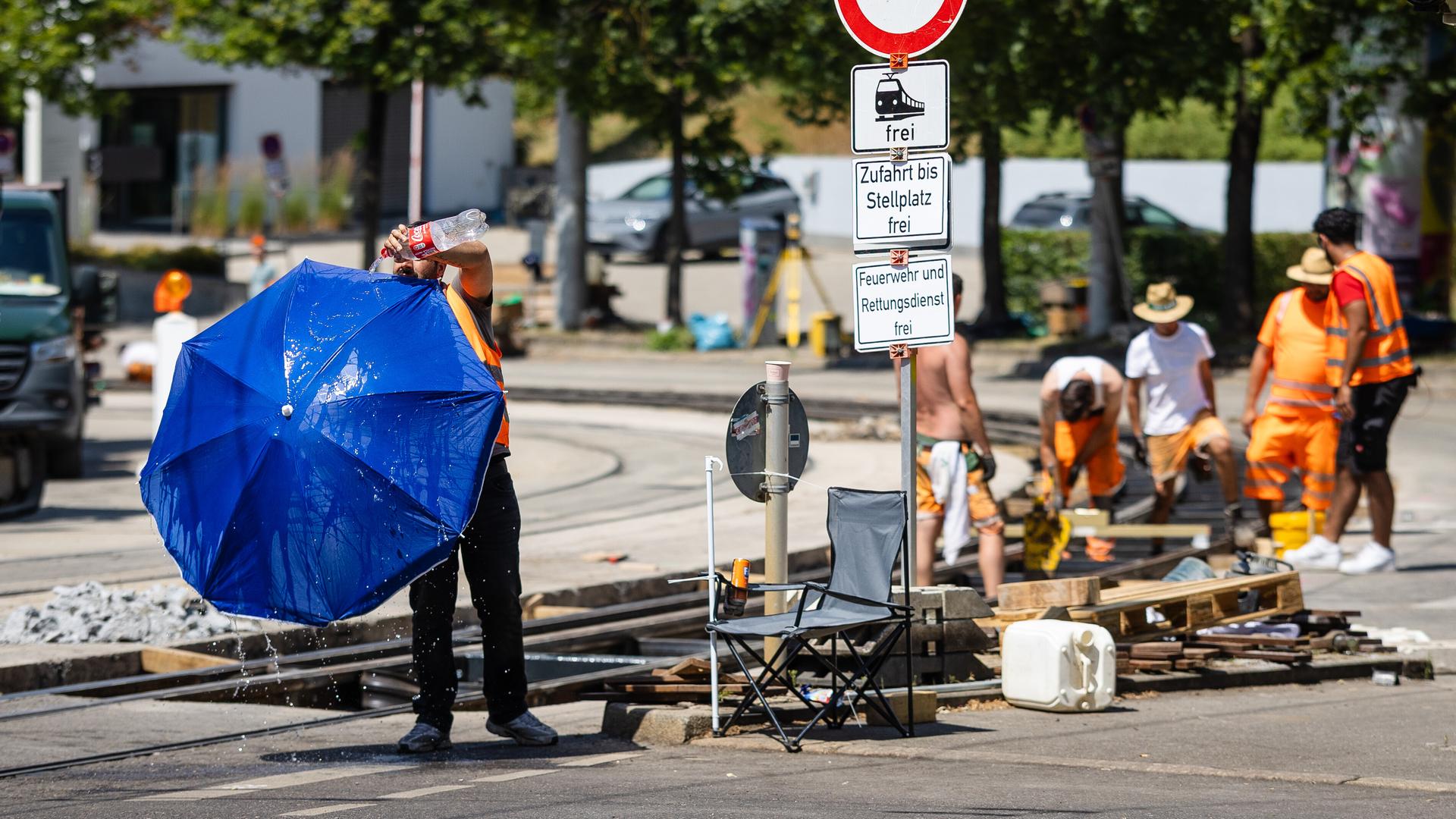 Auf einer Baustelle am Gleisbett stehen mehrere Bauarbeiter. Einer von ihnen gießt Wasser über einen Sonnenschirm. 