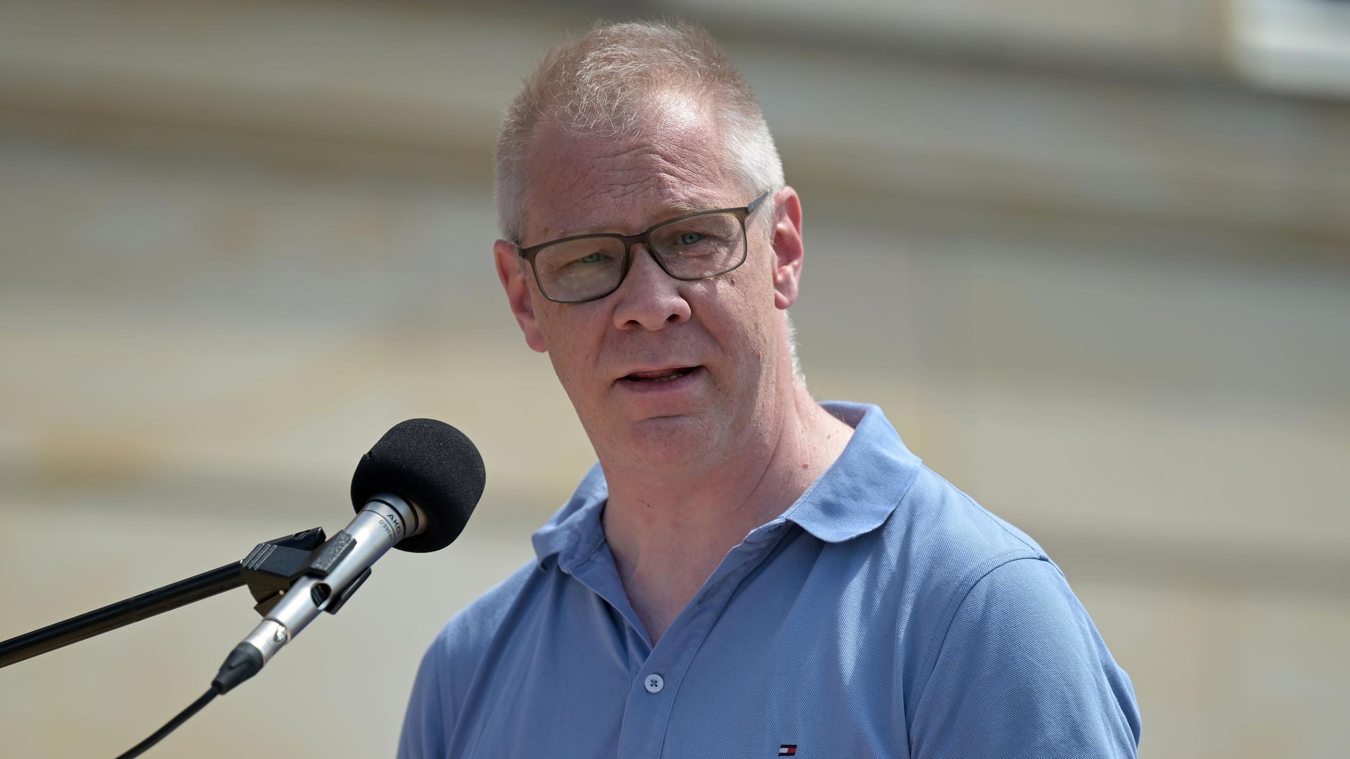 Büttner mit Brille und im hellblauen Hemd steht im Freien vor einem Mikrofon und sagt etwas. Hinter ihm unscharf eine beigefarbene Wand. Büttner mit Brille und im hellblauen Hemd steht im Freien vor einem Mikrofon und sagt etwas. Hinter ihm unscharf eine beigefarbene Wand.