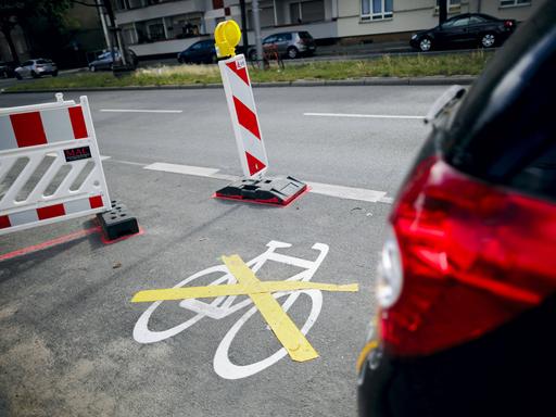 Das Piktogramm fuer einen neuen Radweg wurde mit einer gelben Markierung durchkreuzt. Daneben parkt ein Auto.