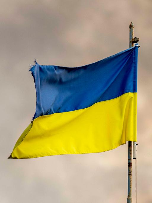 Zu sehen ist die blau-gelbe ukrainische Flagge an einem Fahnenmast vor einem wolkenverhangenen Himmel. Der Fahnenmast ist rostig. Die Flagge etwas zerrissen.