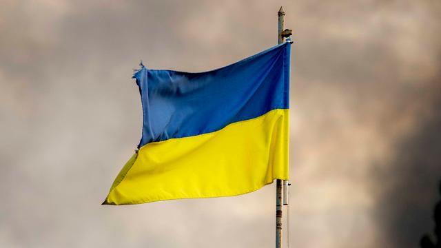 Zu sehen ist die blau-gelbe ukrainische Flagge an einem Fahnenmast vor einem wolkenverhangenen Himmel. Der Fahnenmast ist rostig. Die Flagge etwas zerrissen.