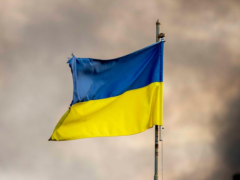 Zu sehen ist die blau-gelbe ukrainische Flagge an einem Fahnenmast vor einem wolkenverhangenen Himmel. Der Fahnenmast ist rostig. Die Flagge etwas zerrissen.