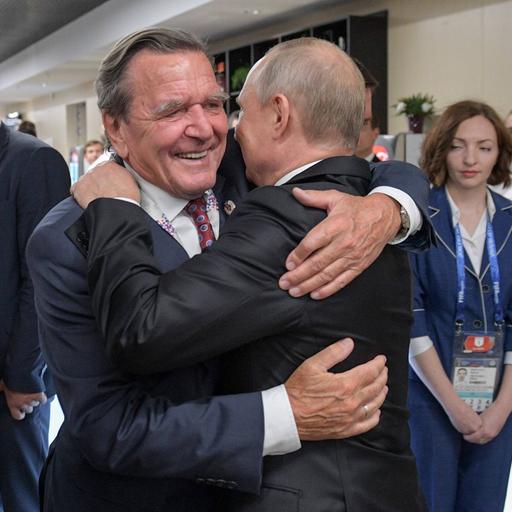 Wladimir Putin und Gerhard Schröder stehen zwischen mehreren Menschen und umarmen sich zur Begrüßung.