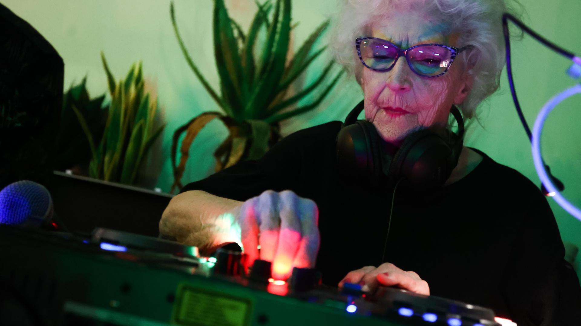 Die 86-jährige DJ Vika steht hinter einem Mischer und legt auf. Sie trägt eine Sonnenbrille, die Wand hinter ihr schimmert grünlich. 