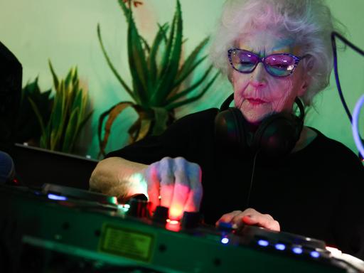 Die 86-jährige DJ Vika steht hinter einem Mischer und legt auf. Sie trägt eine Sonnenbrille, die Wand hinter ihr schimmert grünlich. 