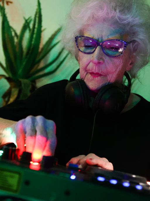 Die 86-jährige DJ Vika steht hinter einem Mischer und legt auf. Sie trägt eine Sonnenbrille, die Wand hinter ihr schimmert grünlich. 