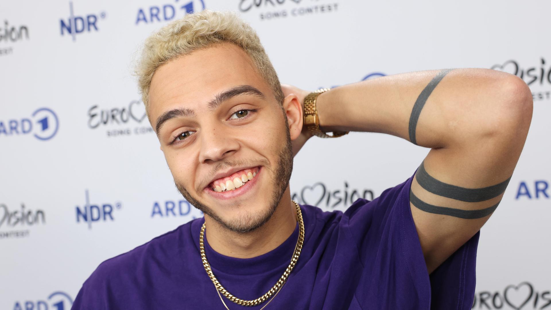 Malik Harris lächelt in die Kamera. Er trägt ein lila T-Shirt. Eine Goldkettte schmückt seinen Hals. Malik Harris lächelt in die Kamera. Er trägt ein lila T-Shirt. Eine Goldkettte schmückt seinen Hals.