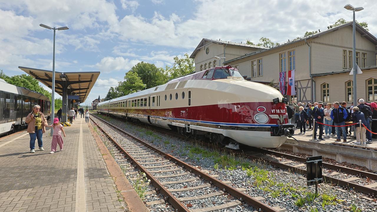 Ein alter Schnellzug. Er ist beige und dunkelrot lackiert. Er steht im Sonnenschein an einem Bahnhof. Auf dem Bahnsteig stehen viele Menschen. 