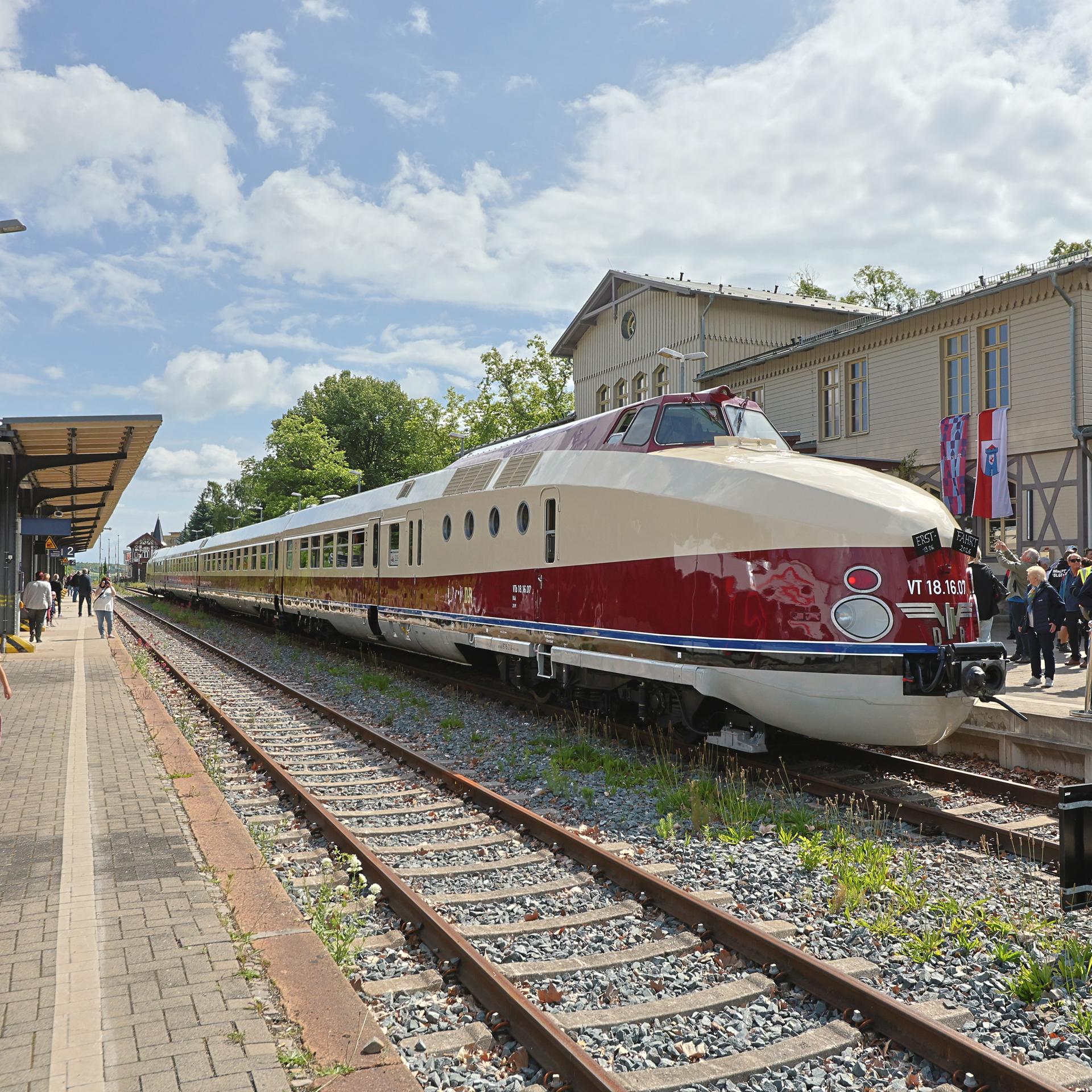 Ein alter Schnellzug. Er ist beige und dunkelrot lackiert. Er steht im Sonnenschein an einem Bahnhof. Auf dem Bahnsteig stehen viele Menschen. 