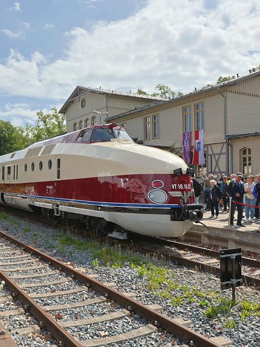Ein alter Schnellzug. Er ist beige und dunkelrot lackiert. Er steht im Sonnenschein an einem Bahnhof. Auf dem Bahnsteig stehen viele Menschen. 
