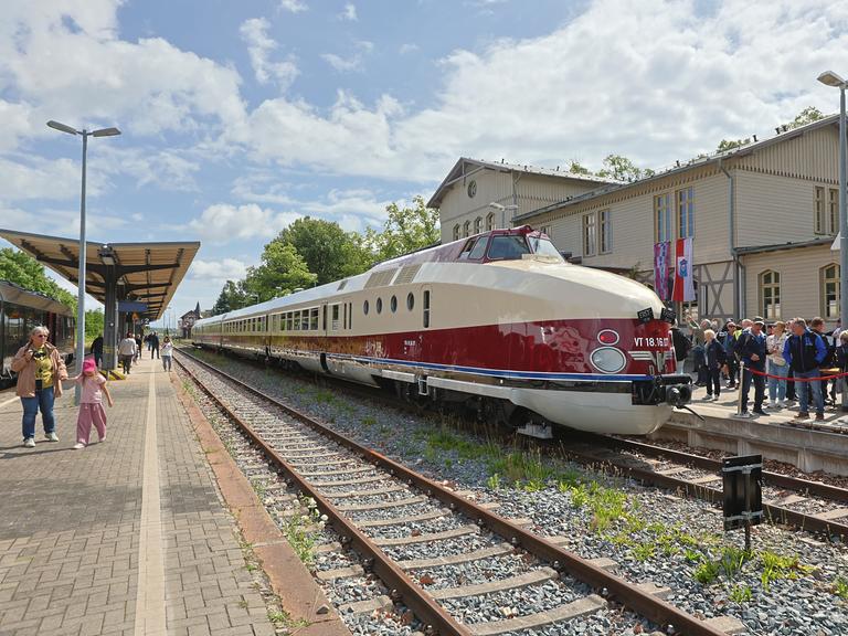 Ein alter Schnellzug. Er ist beige und dunkelrot lackiert. Er steht im Sonnenschein an einem Bahnhof. Auf dem Bahnsteig stehen viele Menschen. 