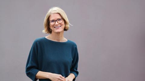 Portrait einer Frau mit blonden, kinnlangen Haaren und Brille, die lächelnd mit geneigtem Kopf in die Kamera blickt.