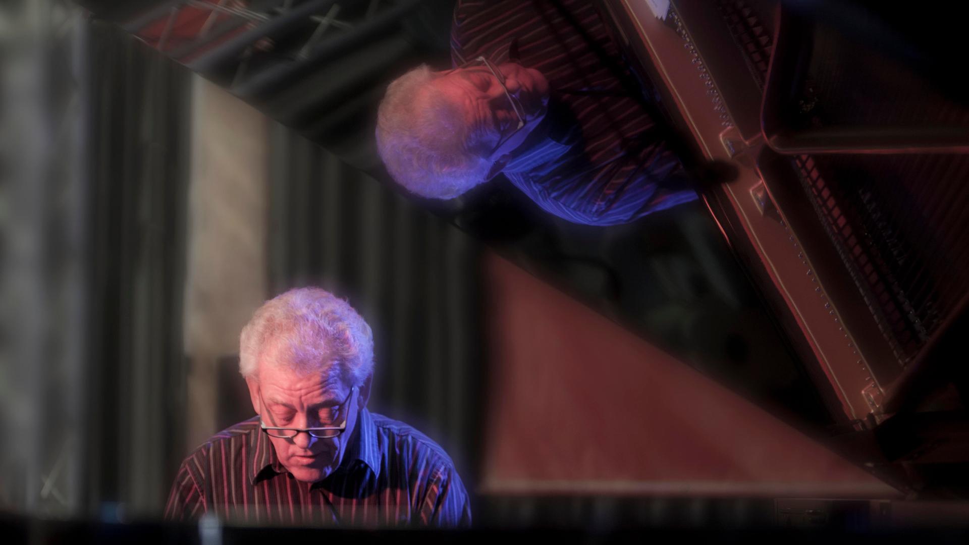 John Taylor sitzt im Jahr 2011 am Flügel und spielt, während er sich im Lack des aufgeklappten Instrumentes spiegelt. 