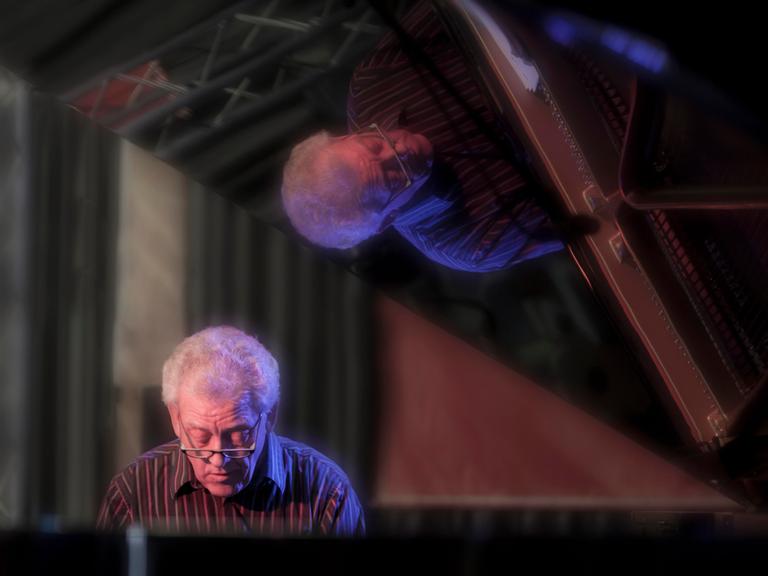 John Taylor sitzt im Jahr 2011 am Flügel und spielt, während er sich im Lack des aufgeklappten Instrumentes spiegelt. 