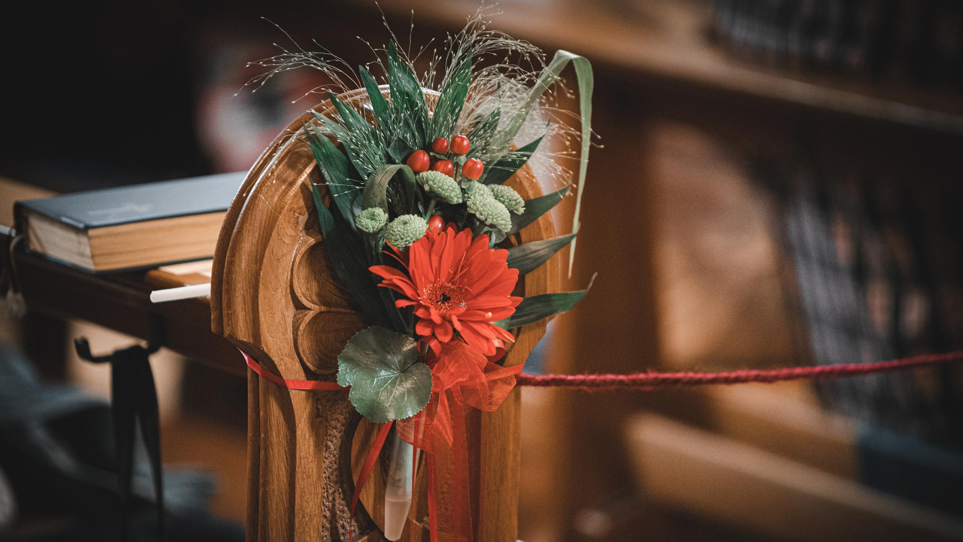 Ein Blumengesteck mit roten Blüten ist am Rand einer Kirchenbank befestigt.