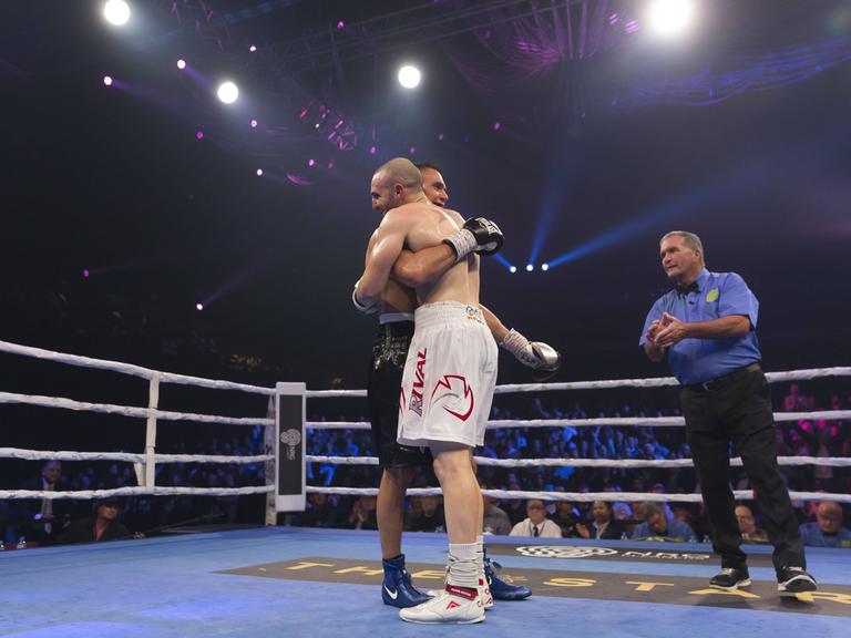 Zwei Boxer - Tim Tszyu and Joel Camilleri - stehen in einem Boxring nach einem Kampf und umarmen sich. Neben ihnen steht ein Ringrichter.